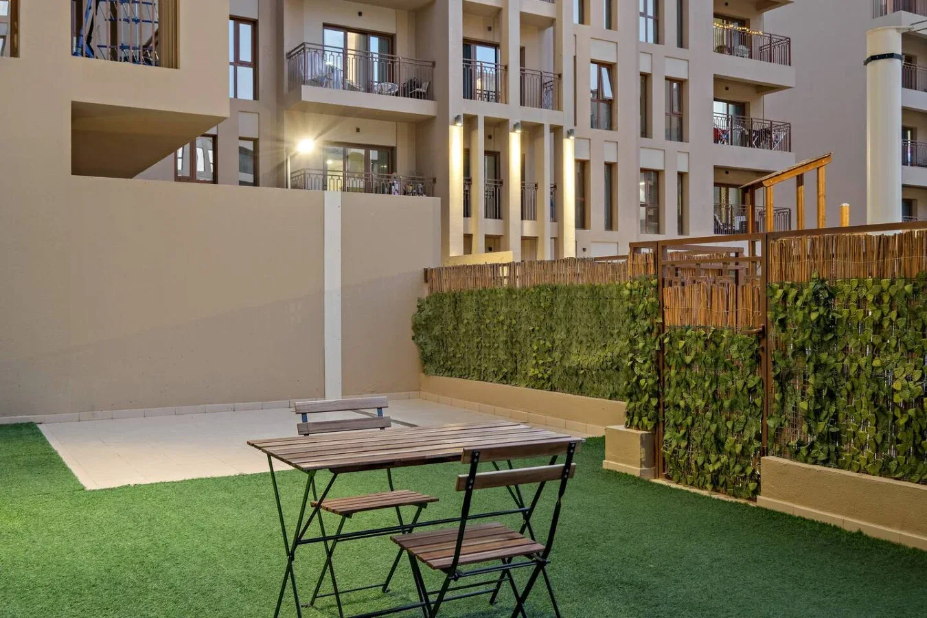 An outdoor seating area on artificial grass sits beside a hedge and apartment building.