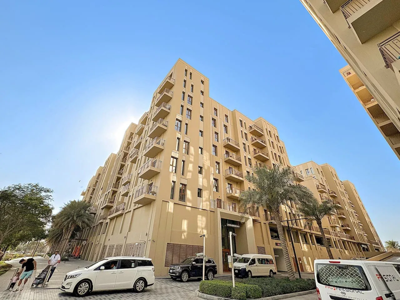 A beige apartment building with balconies stands beside a street under a clear sky.