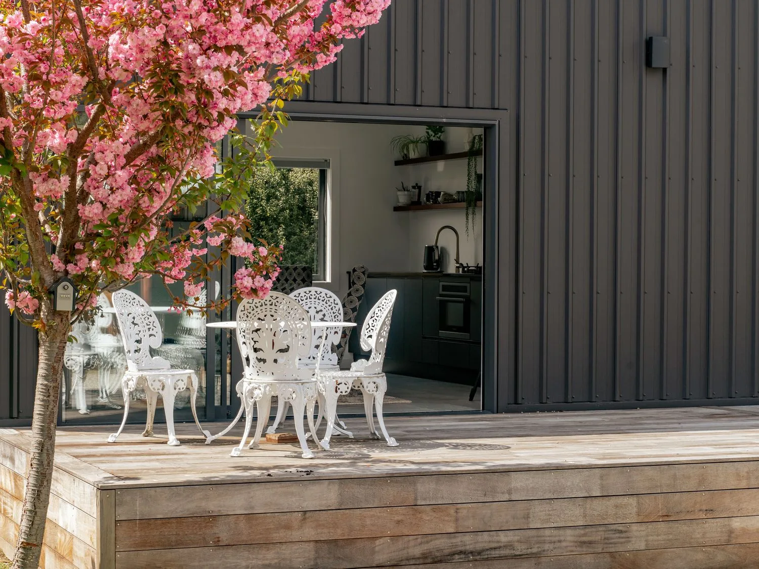 Una terraza de madera con sillas de exterior blancas y una mesa junto a un árbol de flores rosas y un edificio negro en Caledonia - Earnscleugh Holiday Home, en Earnscleugh