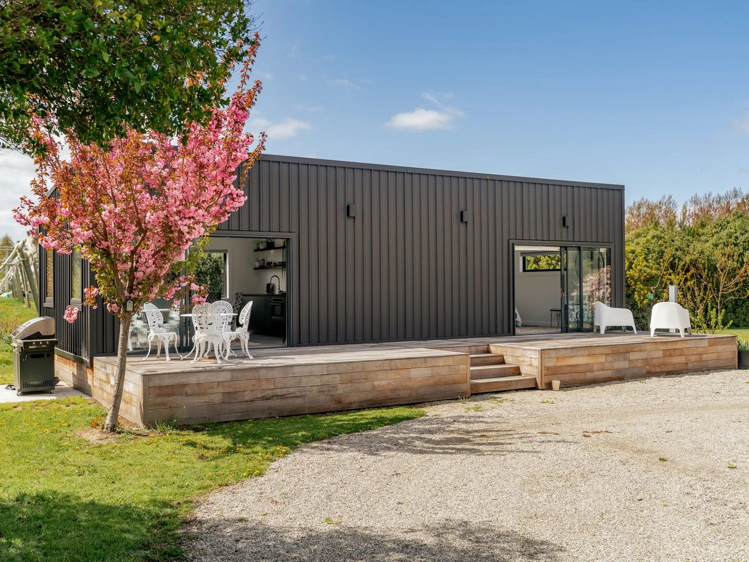 Una casa moderna oscura se encuentra junto a una terraza elevada de madera, asientos al aire libre y un árbol en flor.