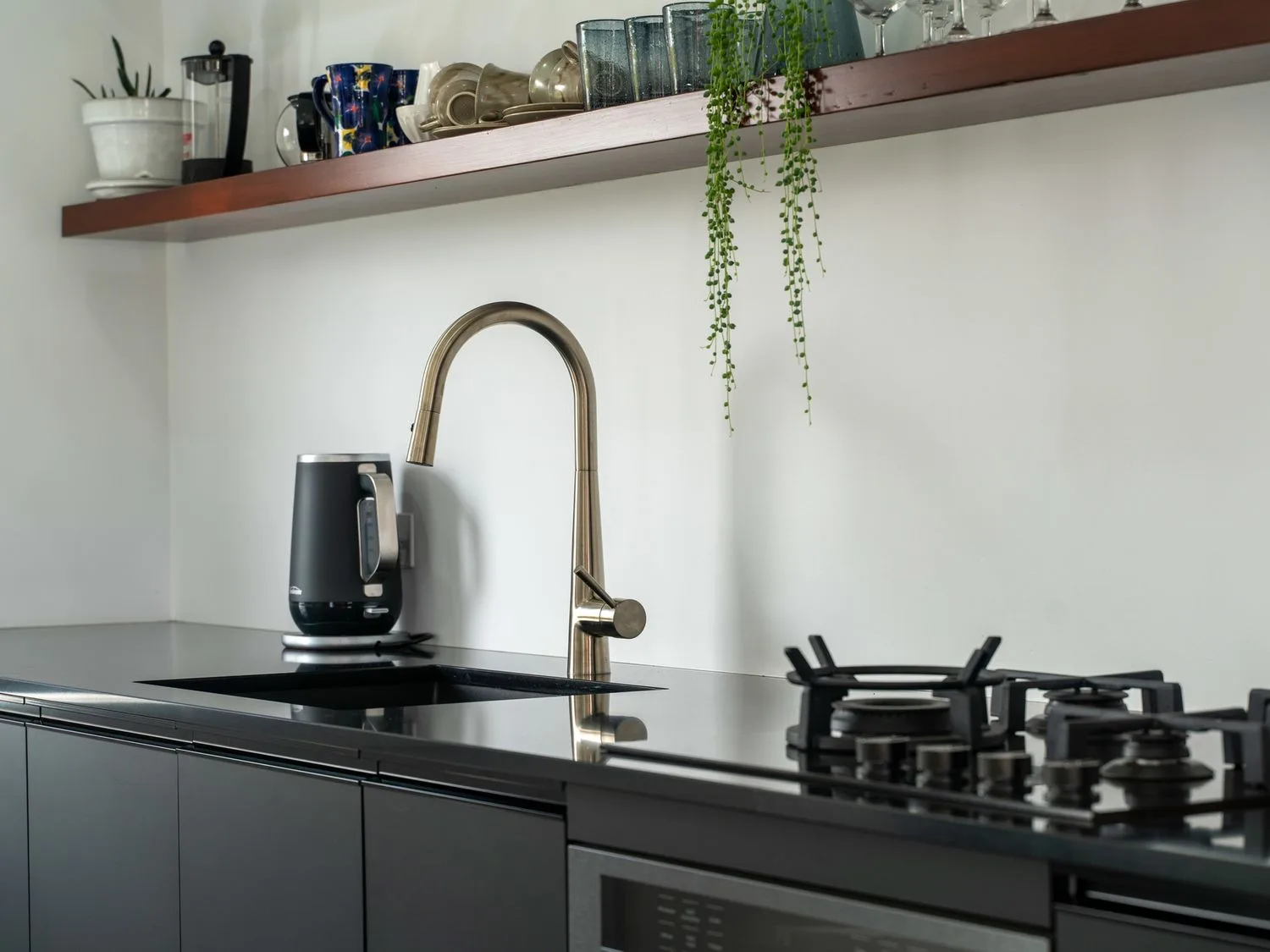 Una encimera de cocina con grifo, hervidor eléctrico, quemadores y un estante abierto encima.