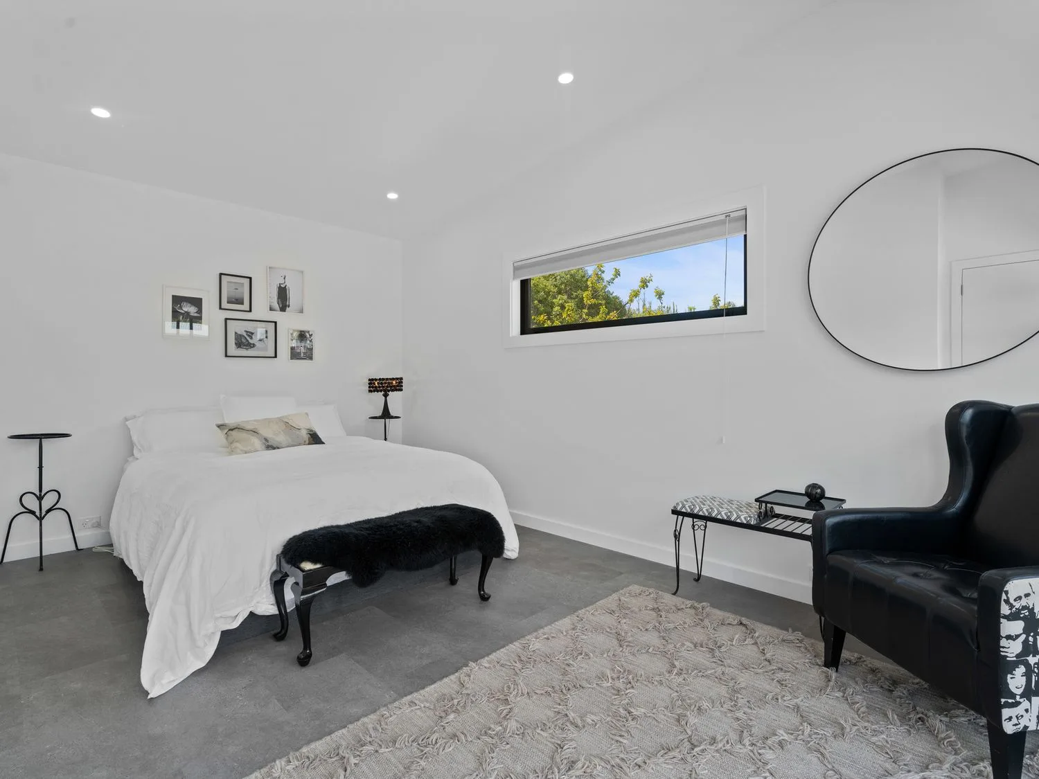 Un dormitorio con ropa de cama blanca, un banco oscuro, cuadros enmarcados y un espejo redondo en la pared.