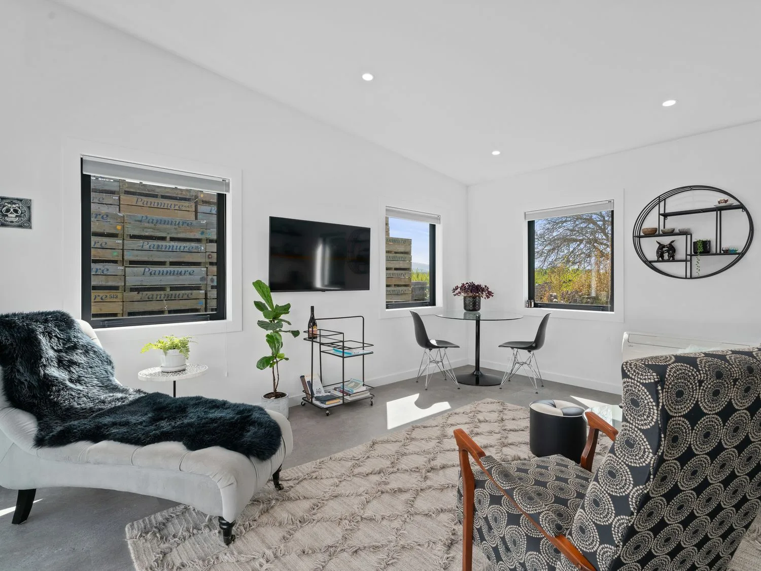 A bright living room with a wall-mounted television, seating, and windows.