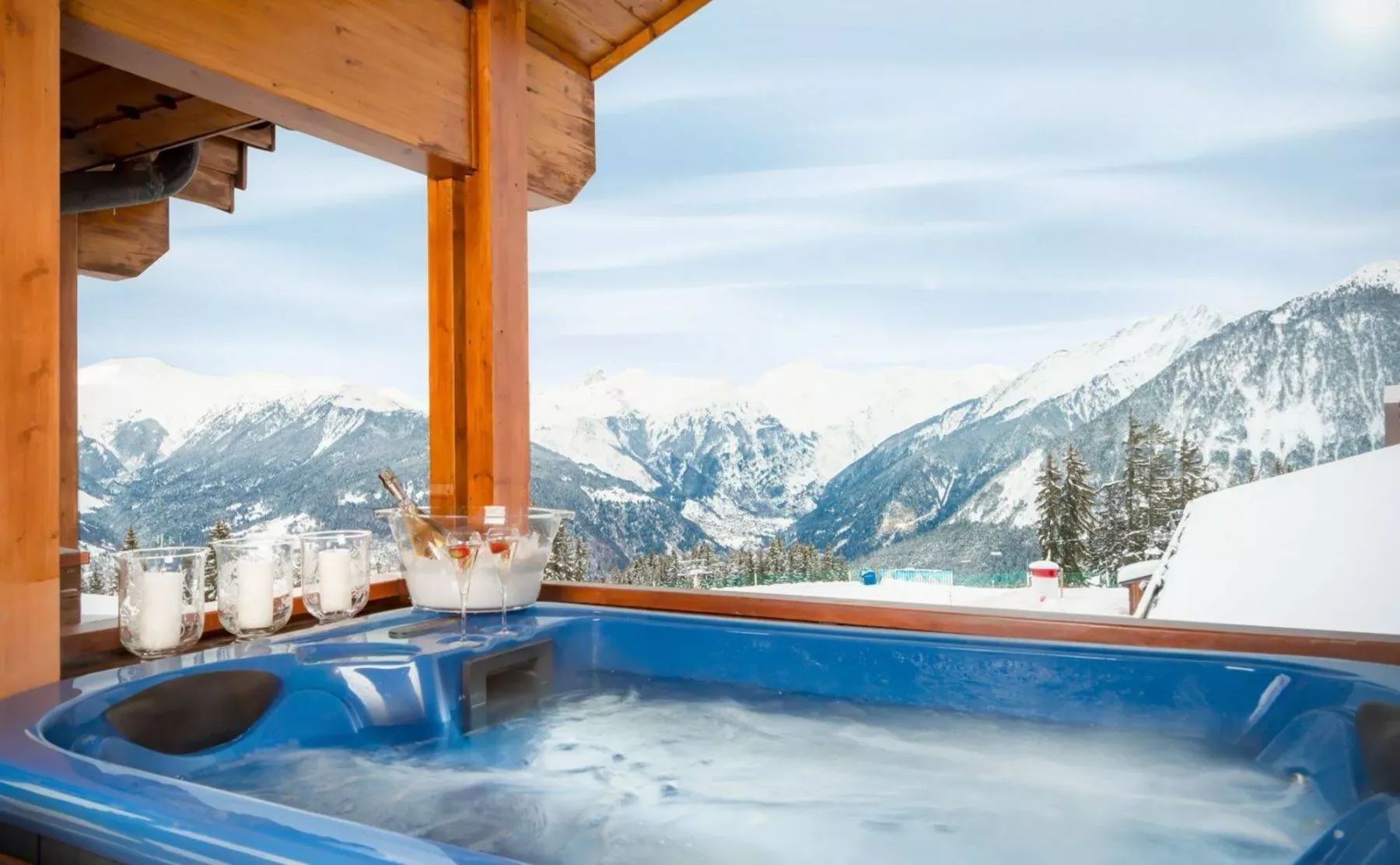 An outdoor hot tub sits beneath a wooden shelter, overlooking snow-covered mountains.