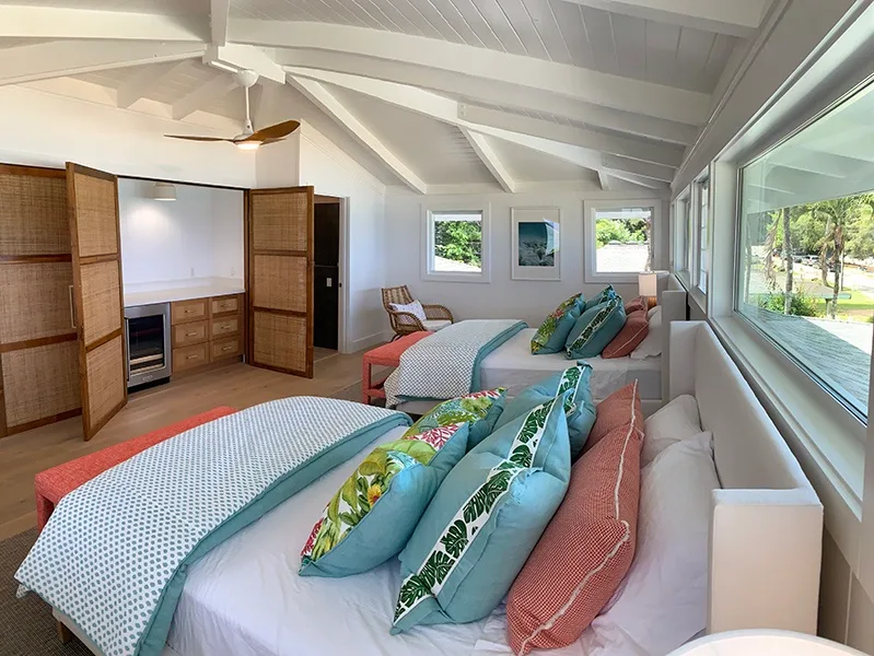 Un dormitorio luminoso con dos camas, almohadas coloridas y ventanas grandes.