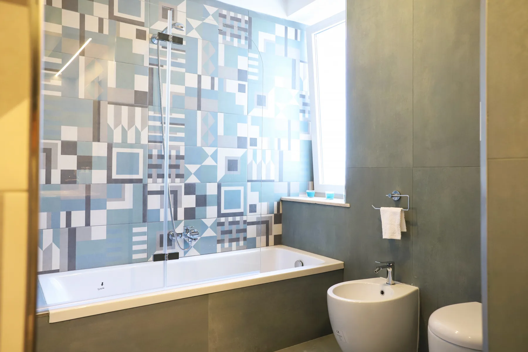 A bathroom with a bathtub, patterned wall tiles, and a bright window.