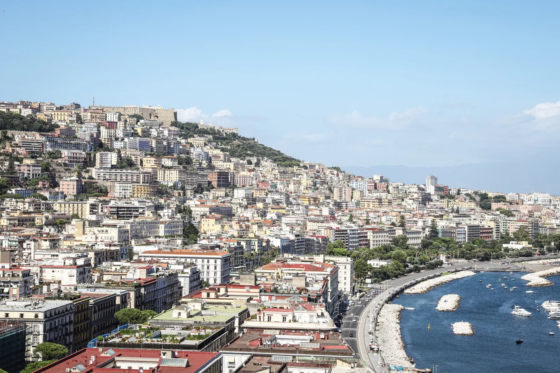 A coastal cityscape shows densely packed buildings climbing a hillside beside a curved waterfront.