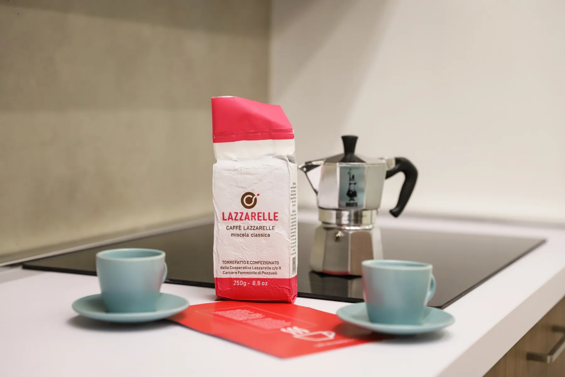 A kitchen counter displays a coffee bag, stovetop espresso maker, and two cups.