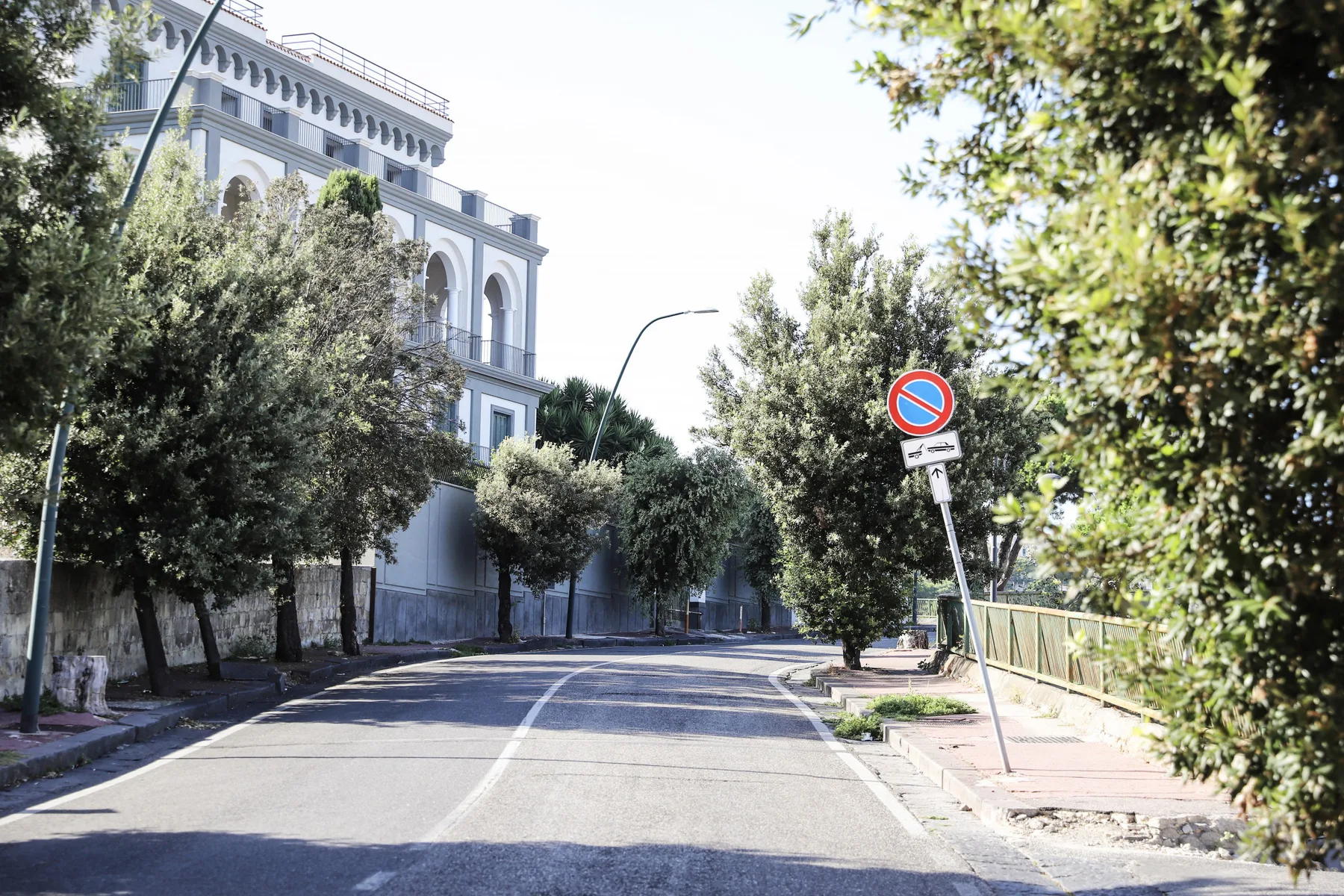 A quiet street curves past a large building lined with trees.
