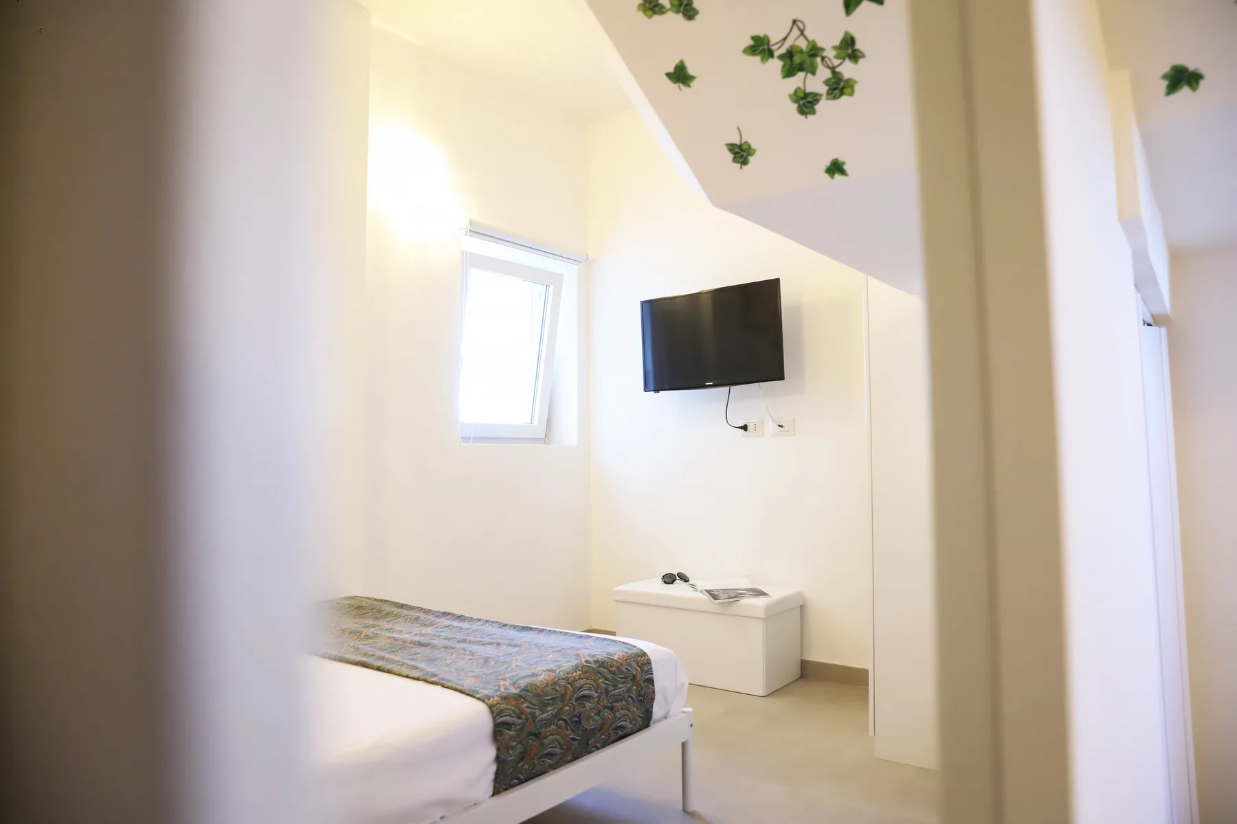 A bright bedroom with a bed, a wall-mounted television, and a small cabinet.