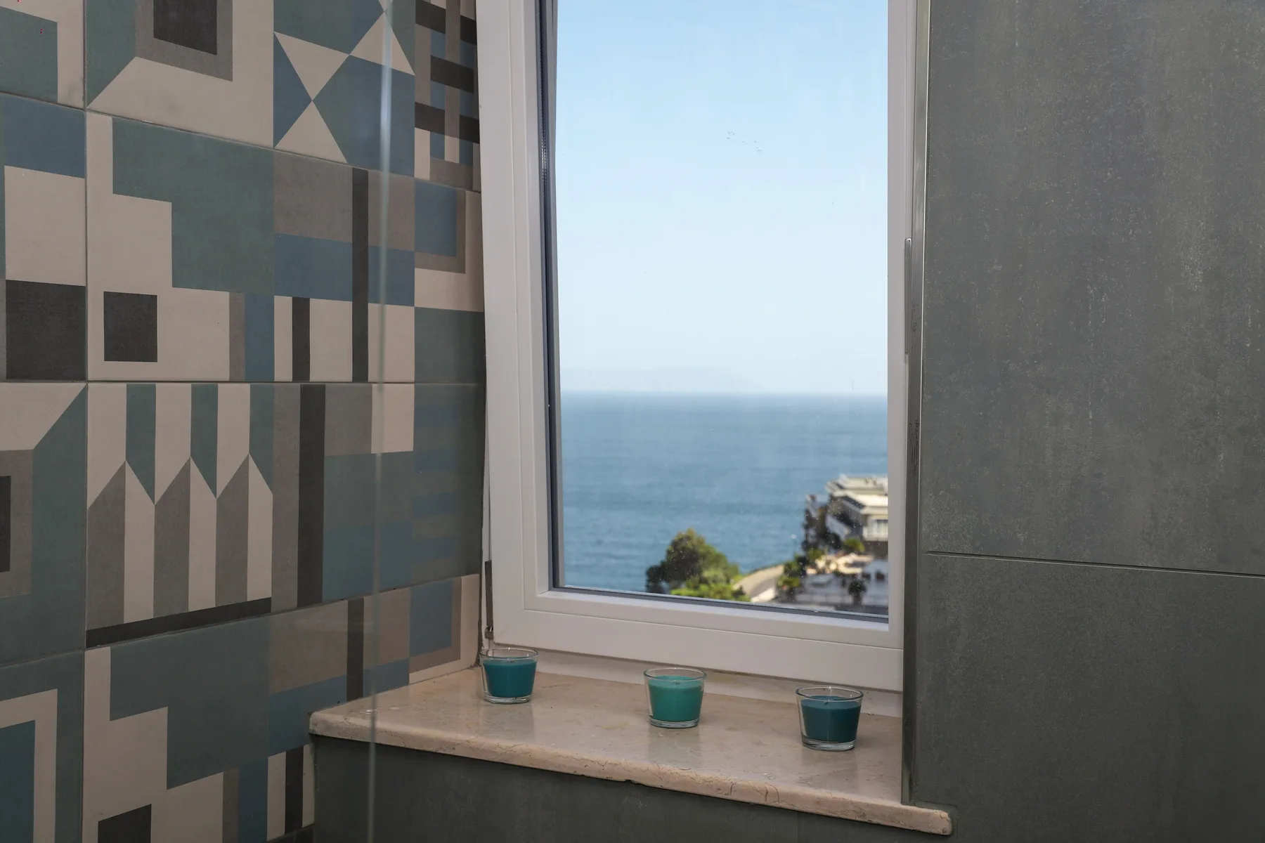 A bathroom window overlooks the sea, with patterned tiles beside it.