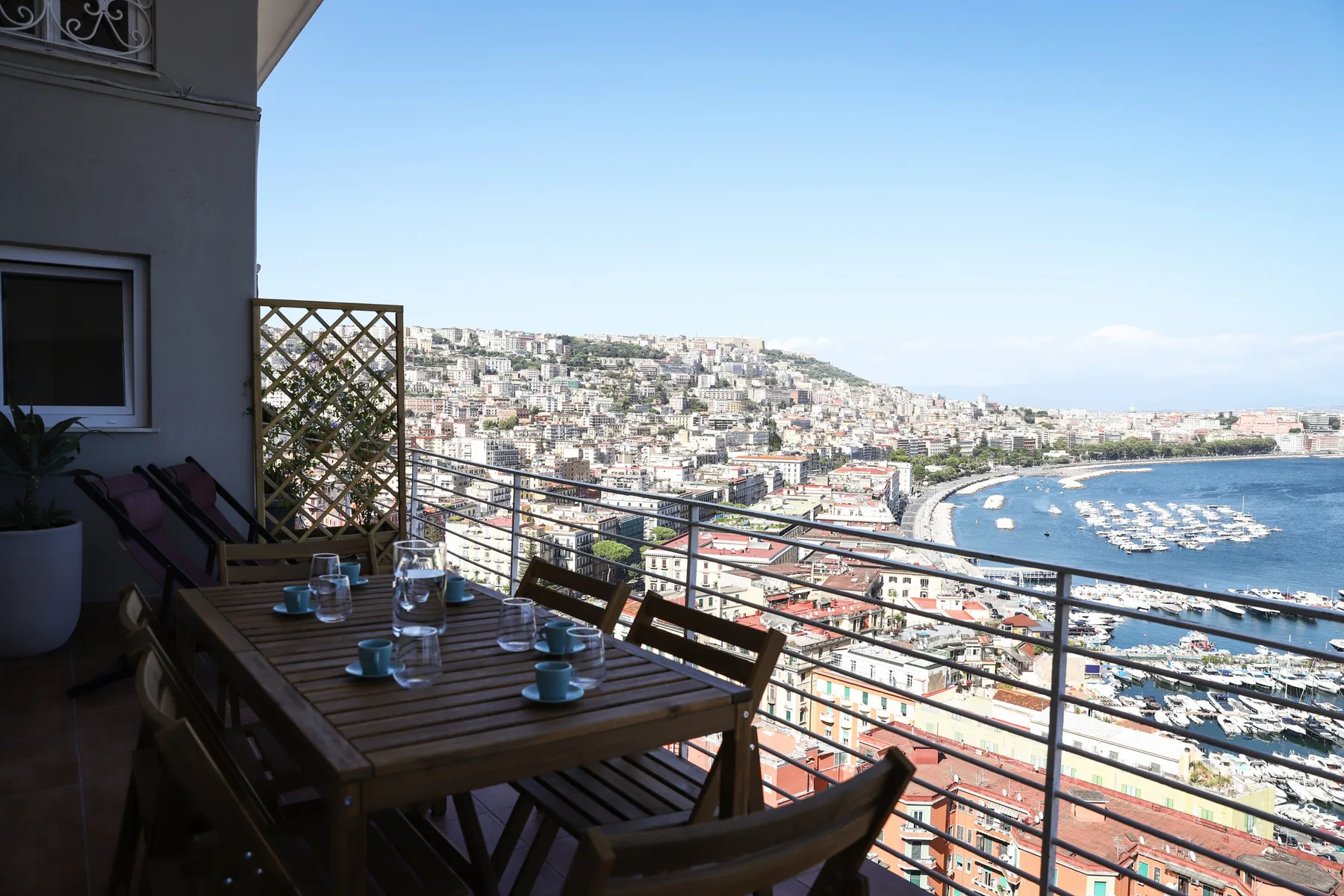 A balcony dining area overlooks a coastal city and a curved harbor.