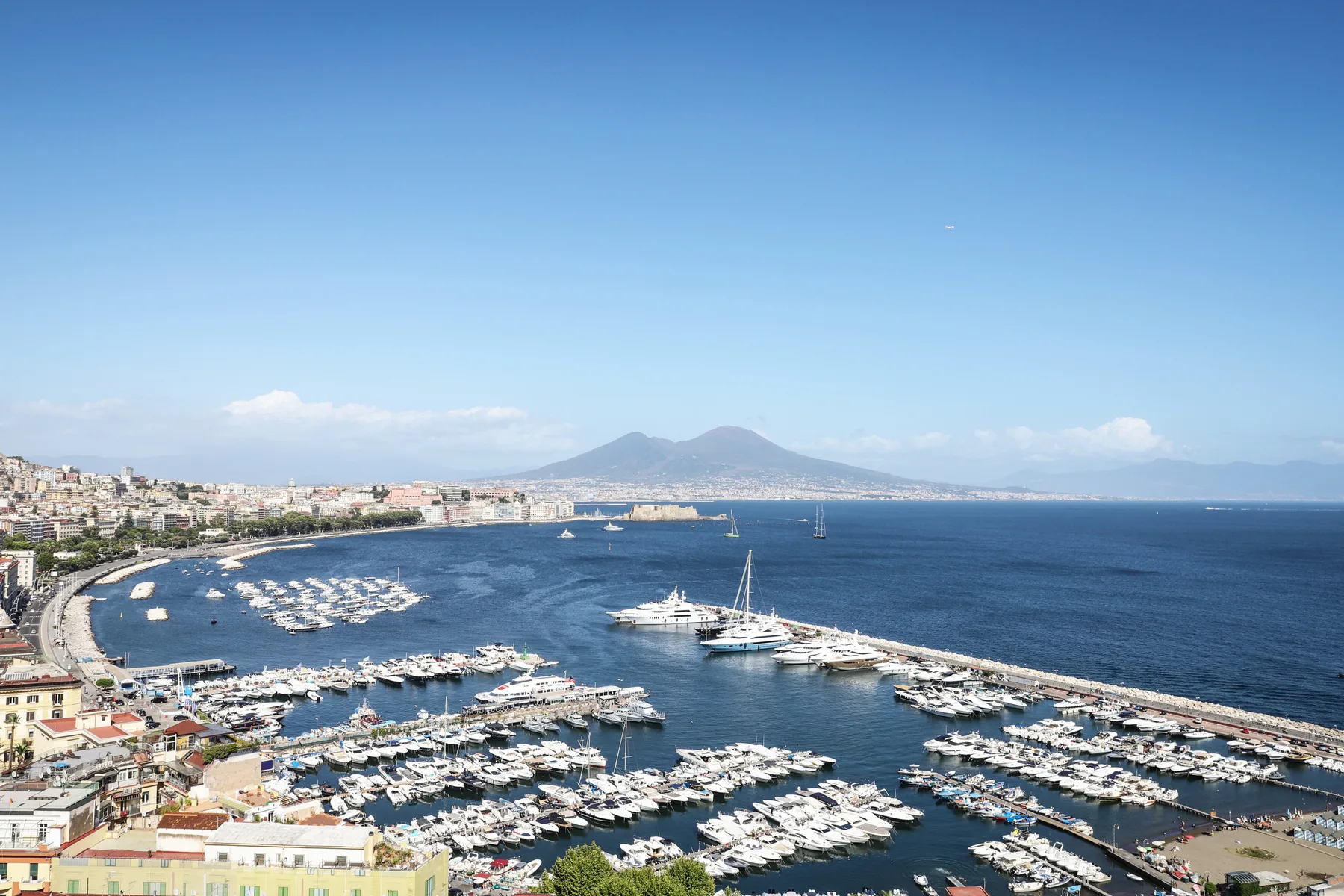 A coastal harbor filled with boats, with a curved shoreline and distant mountains.