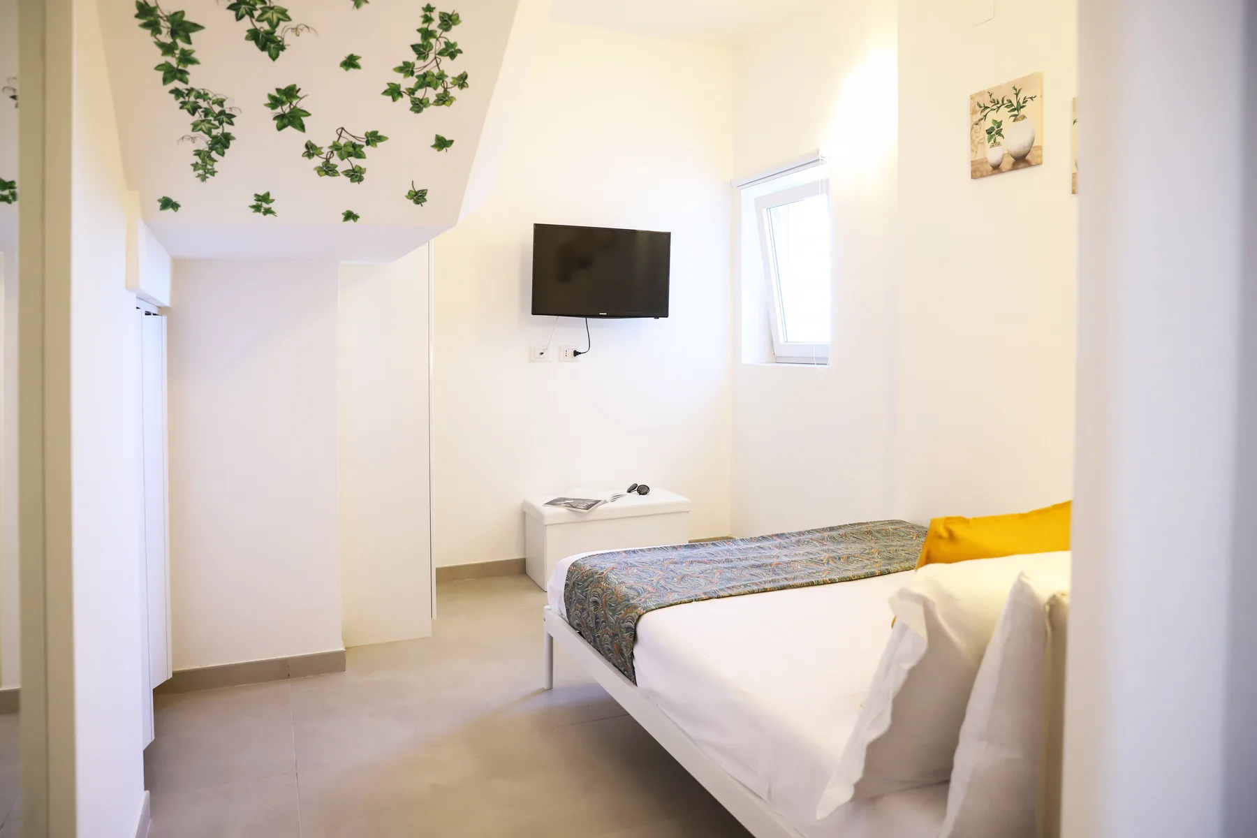 A bright bedroom with a bed, wall-mounted television, and small window.