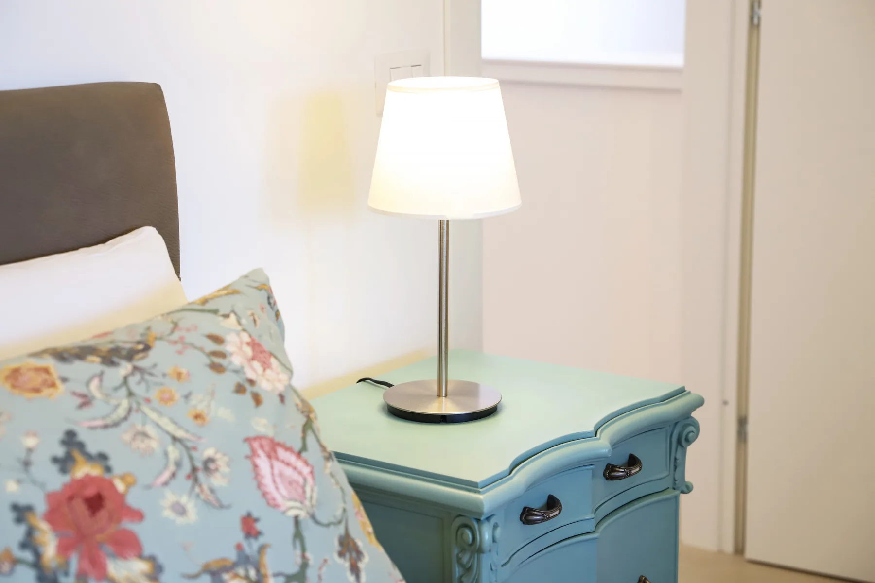 A bedroom scene with a bed beside a blue bedside table and lit lamp.
