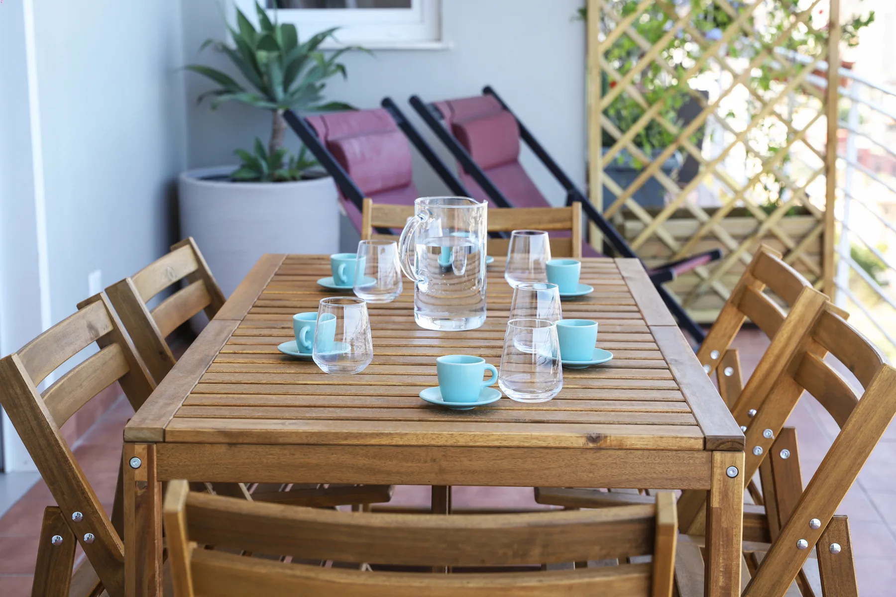 An outdoor dining table is set with glasses and blue cups beside wooden chairs.