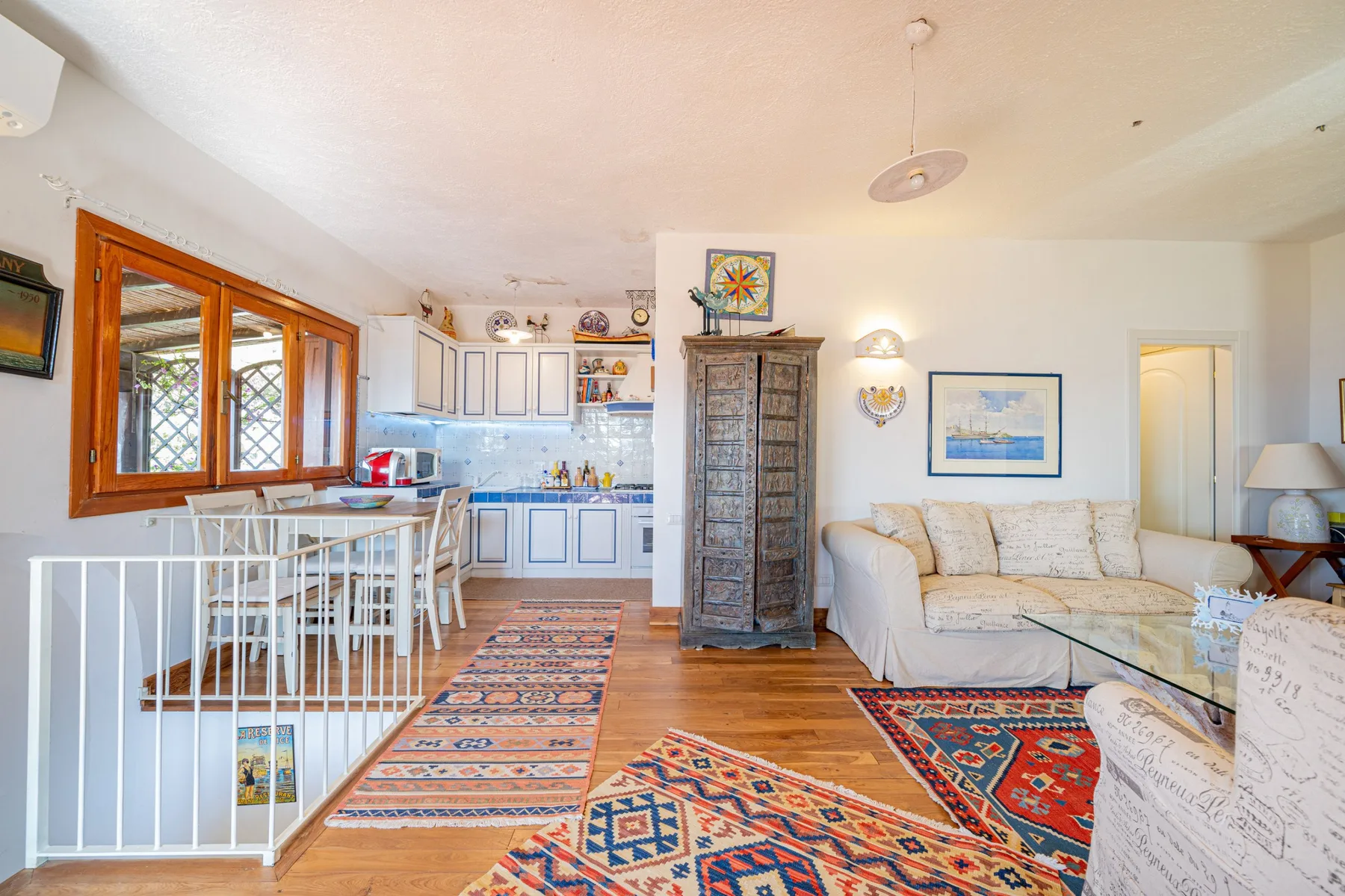 Una luminosa sala de estar y cocina con asientos blancos, gabinetes de madera y alfombras coloridas.