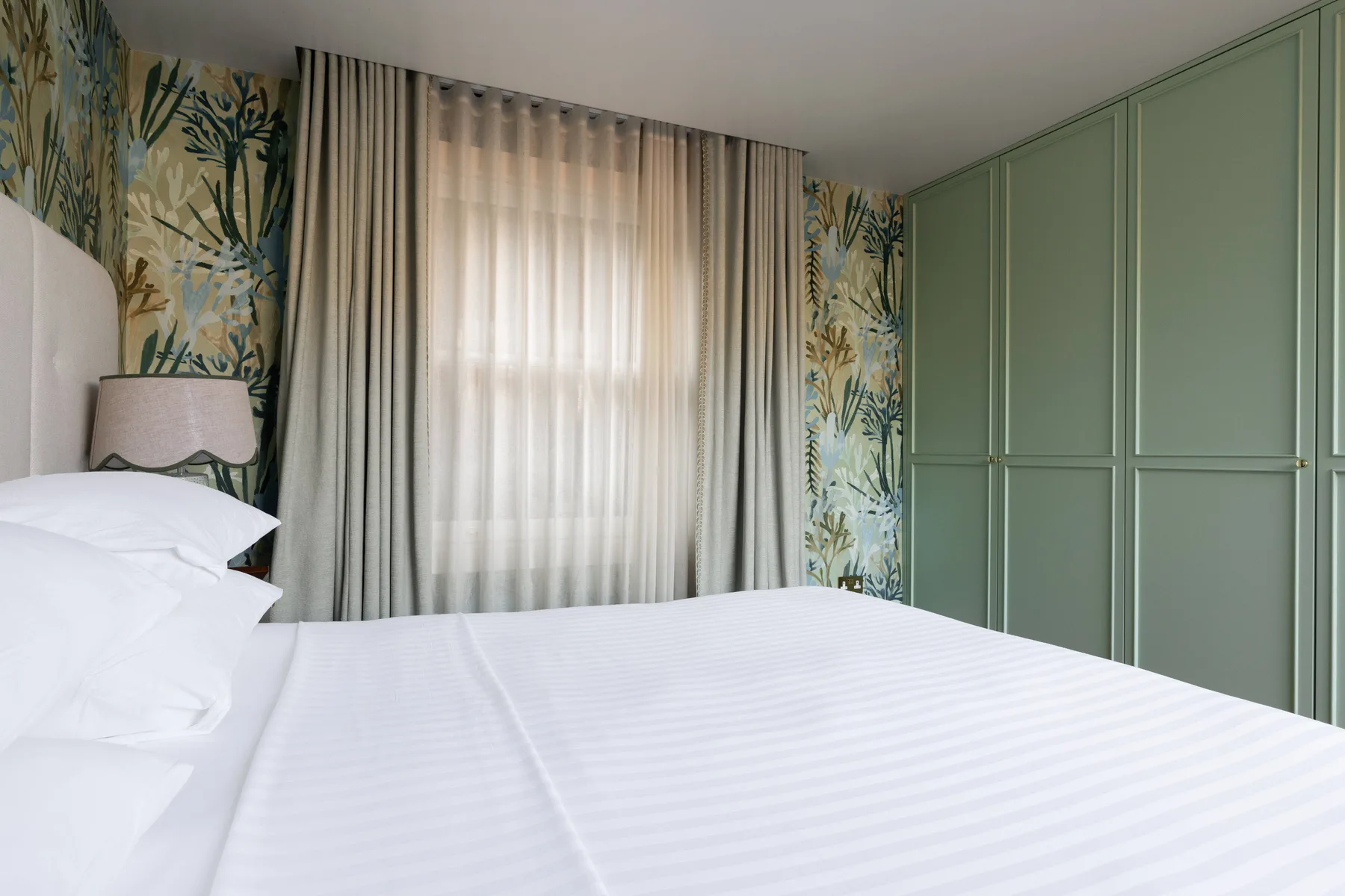 A bedroom with a neatly made bed, patterned wallpaper, and green built-in cabinets.