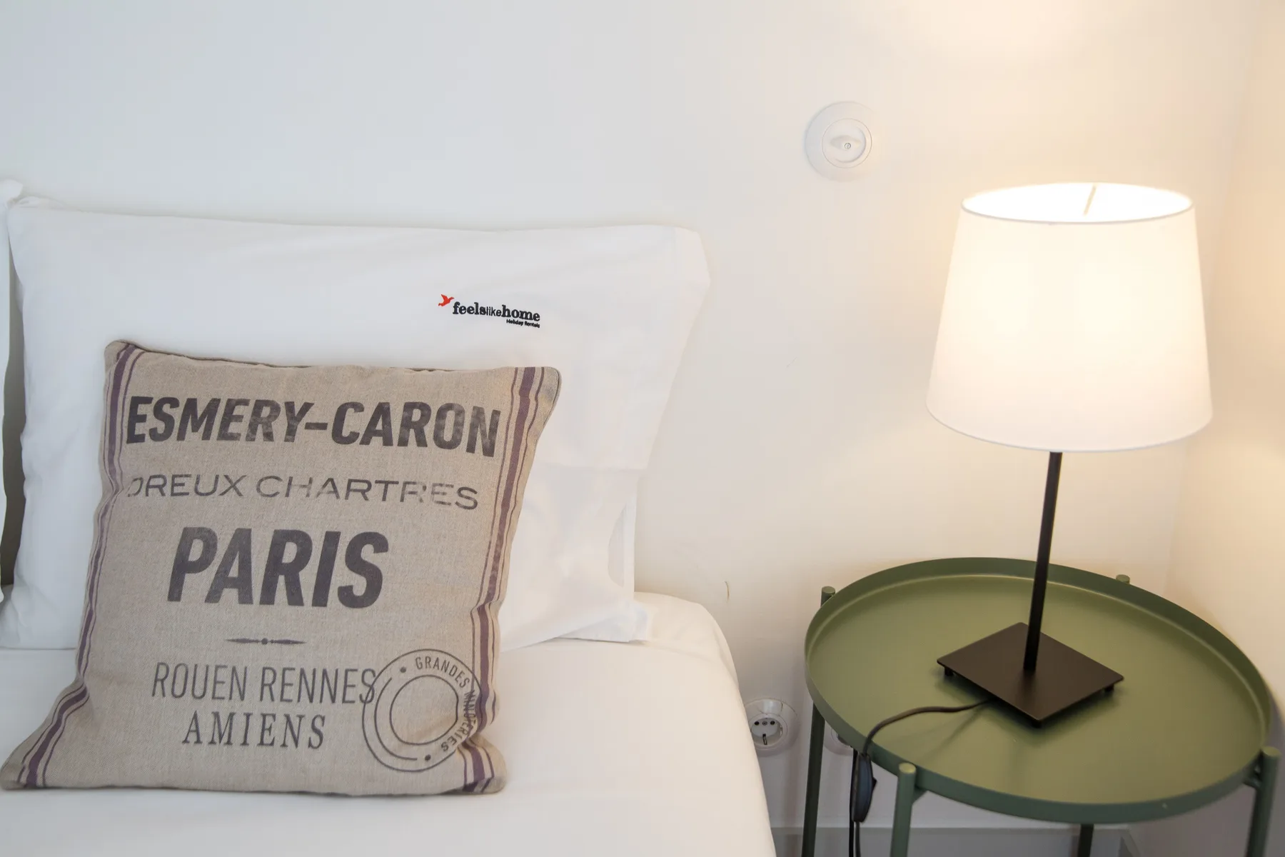 A bed with a printed decorative pillow sits beside a lit table lamp.