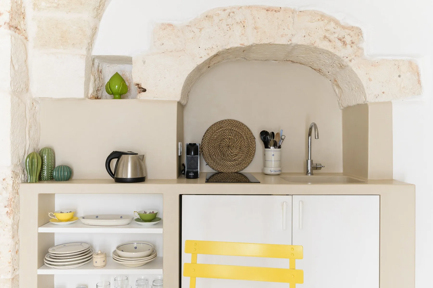 A kitchen counter with a sink, kettle, and open shelves of dishes.
