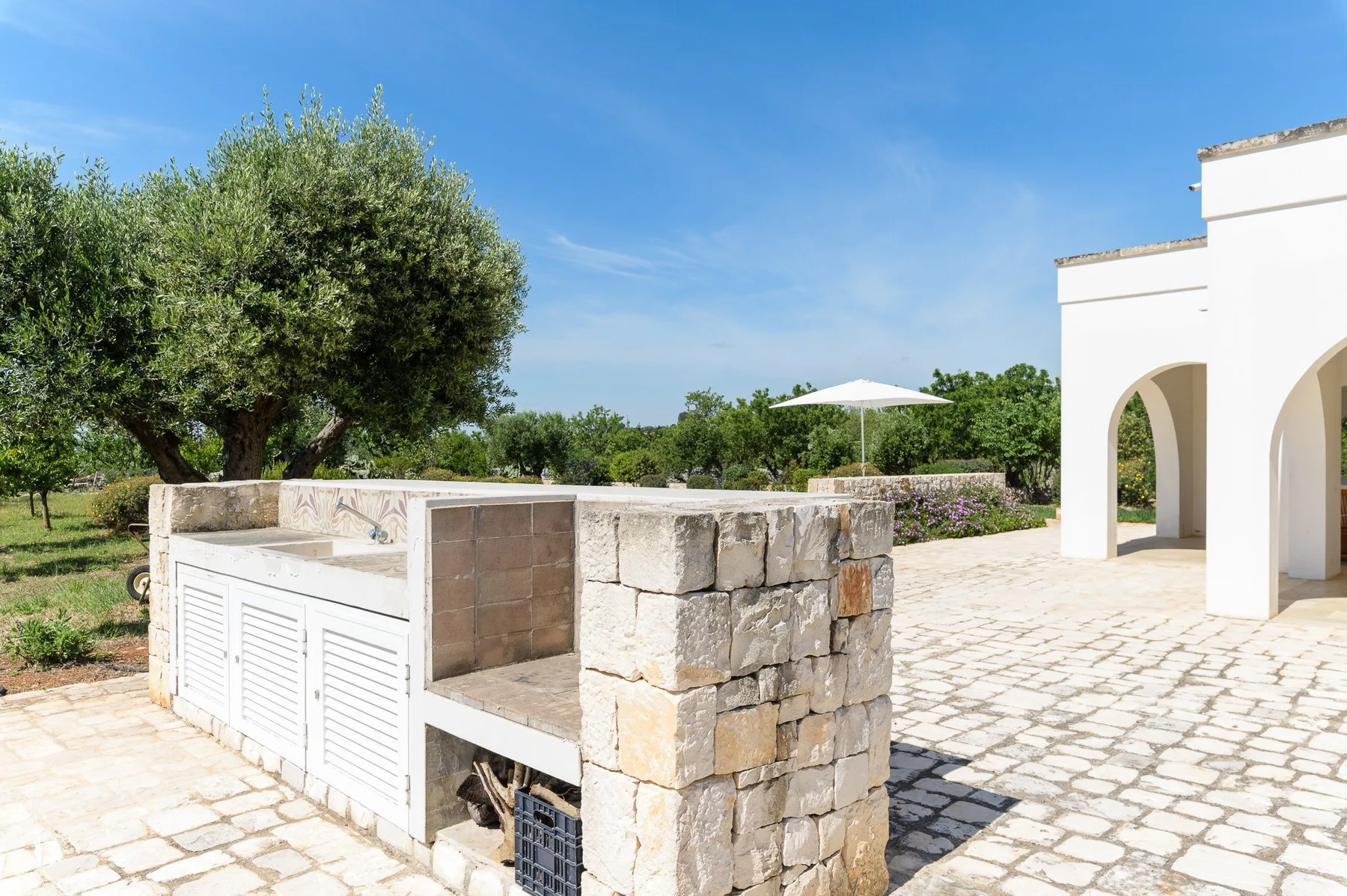 Un patio de piedra soleado con una encimera integrada y un edificio blanco con arcos.