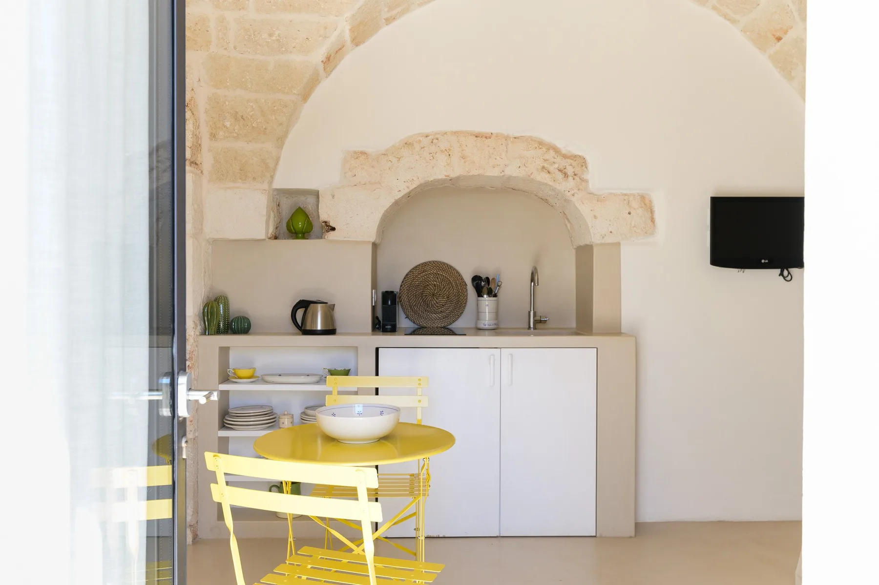 Un comedor luminoso con una mesa amarilla y estantes empotrados bajo un arco de piedra.