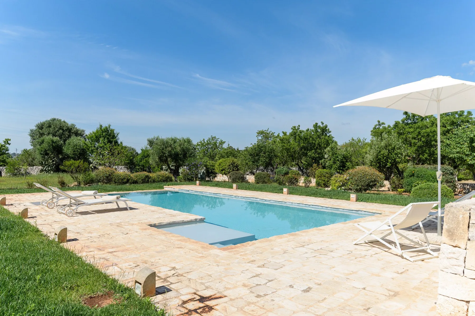 Una piscina al aire libre está rodeada por un patio pavimentado y un jardín.