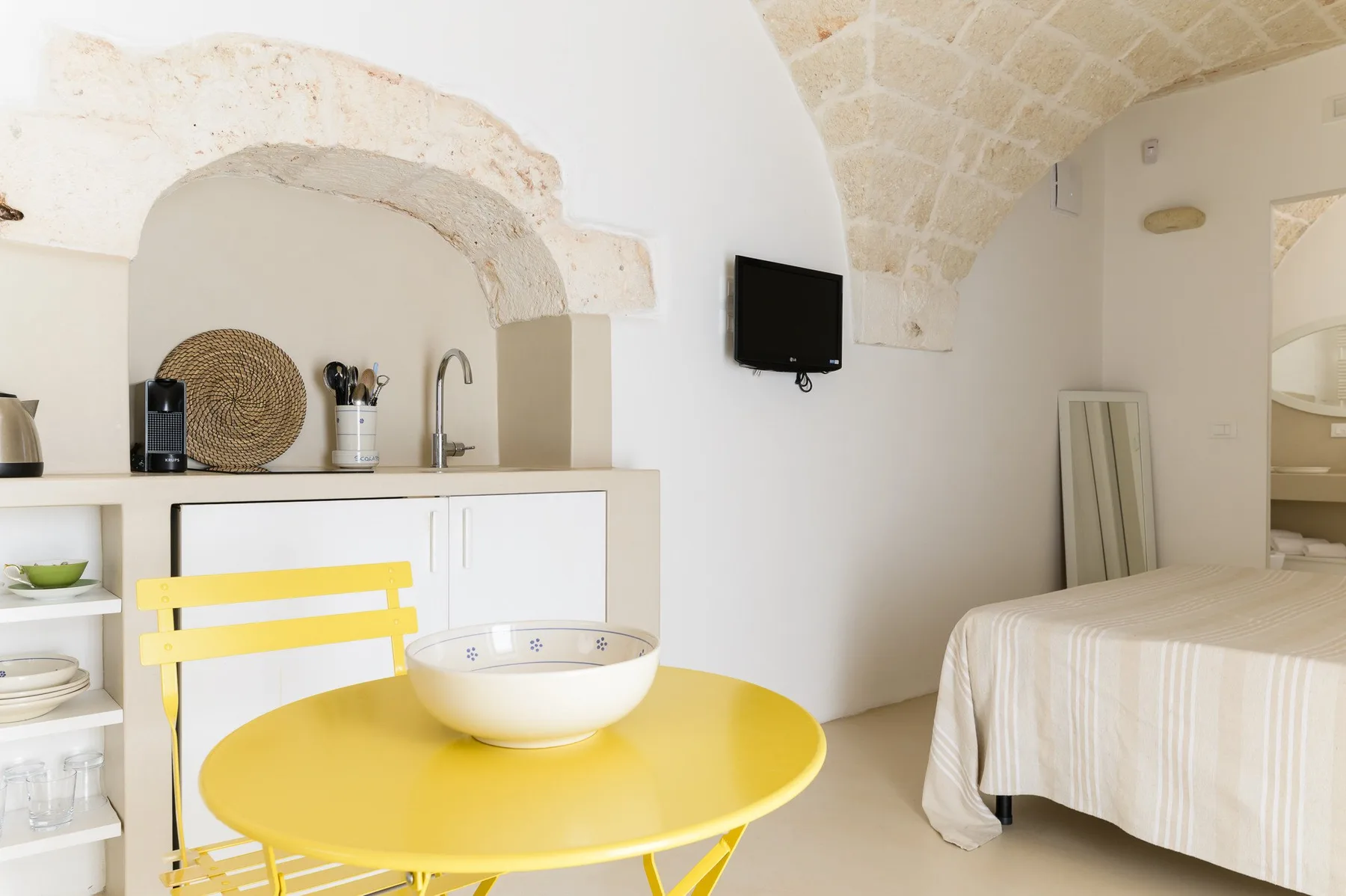 A bedroom with a compact kitchenette, yellow table, and bed beneath a vaulted ceiling.