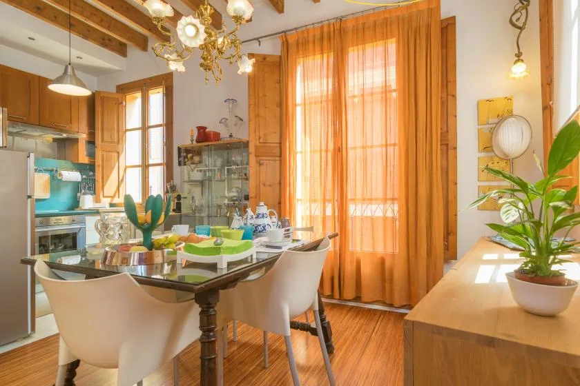 Comedor con la mesa puesta, cortinas naranjas y la cocina al fondo.