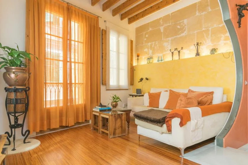 Un dormitorio con una cama blanca, detalles naranjas y ventanas altas con cortinas.