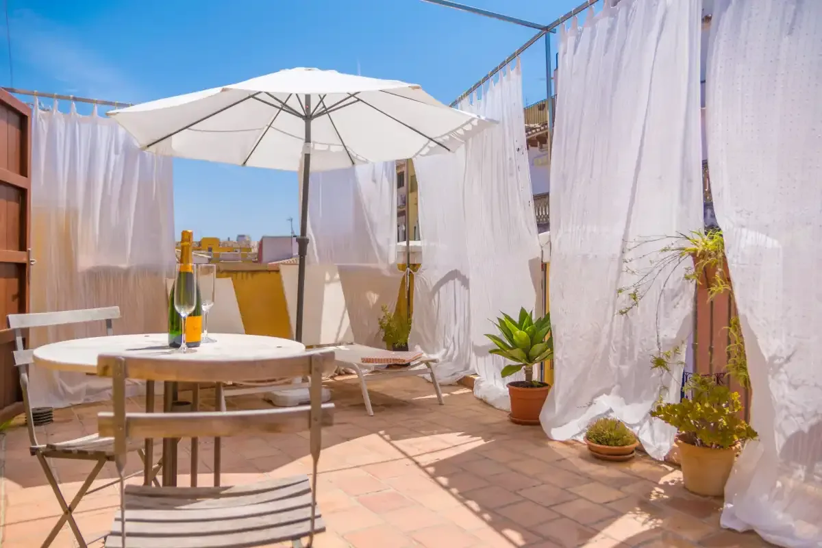 A sunny outdoor terrace has a table, seating, and a large umbrella.