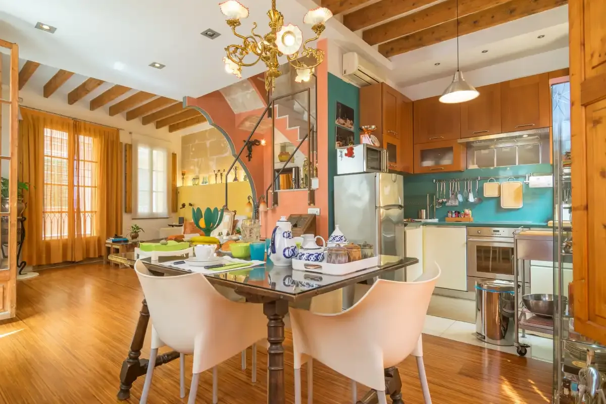 A dining area opens onto a kitchen with wooden cabinets and a turquoise backsplash.