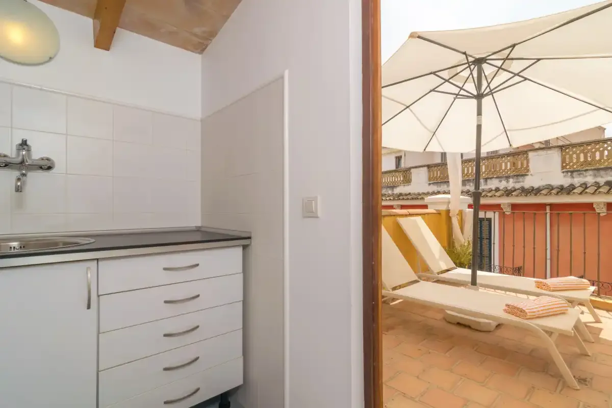 A kitchenette opens onto a sunny terrace with lounge seating beneath an umbrella.