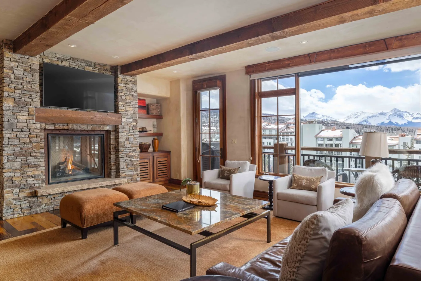 A living room with a stone fireplace and seating beside large windows overlooking snowy mountains.