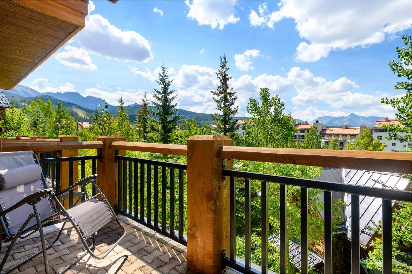 A balcony with seating overlooks trees, nearby buildings, and distant mountains.