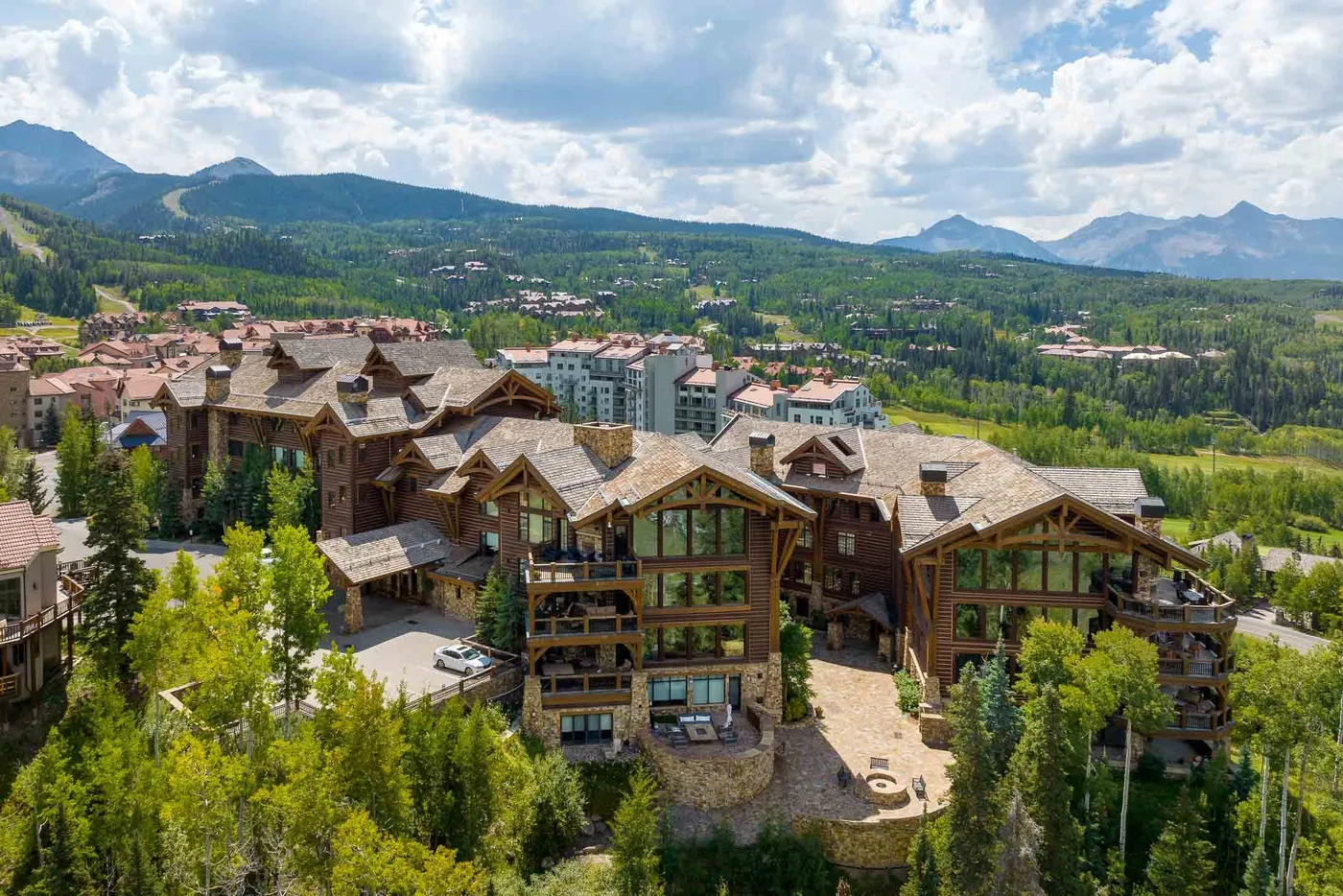 A mountain lodge complex sits among trees, with a green valley and mountain ridges beyond.