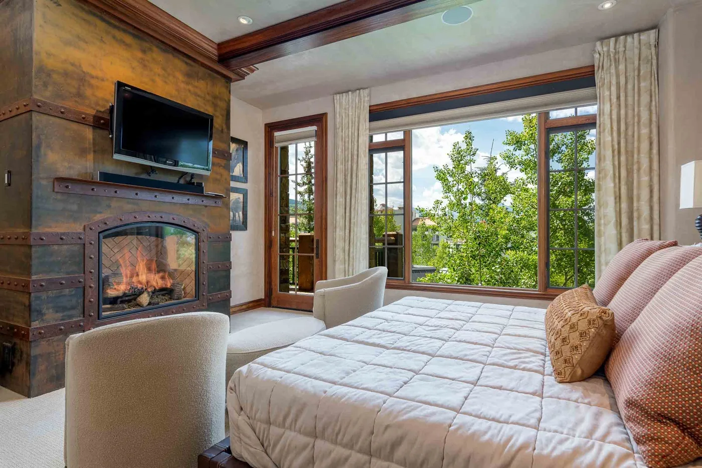 A bedroom with a large bed beside broad windows overlooking leafy trees.