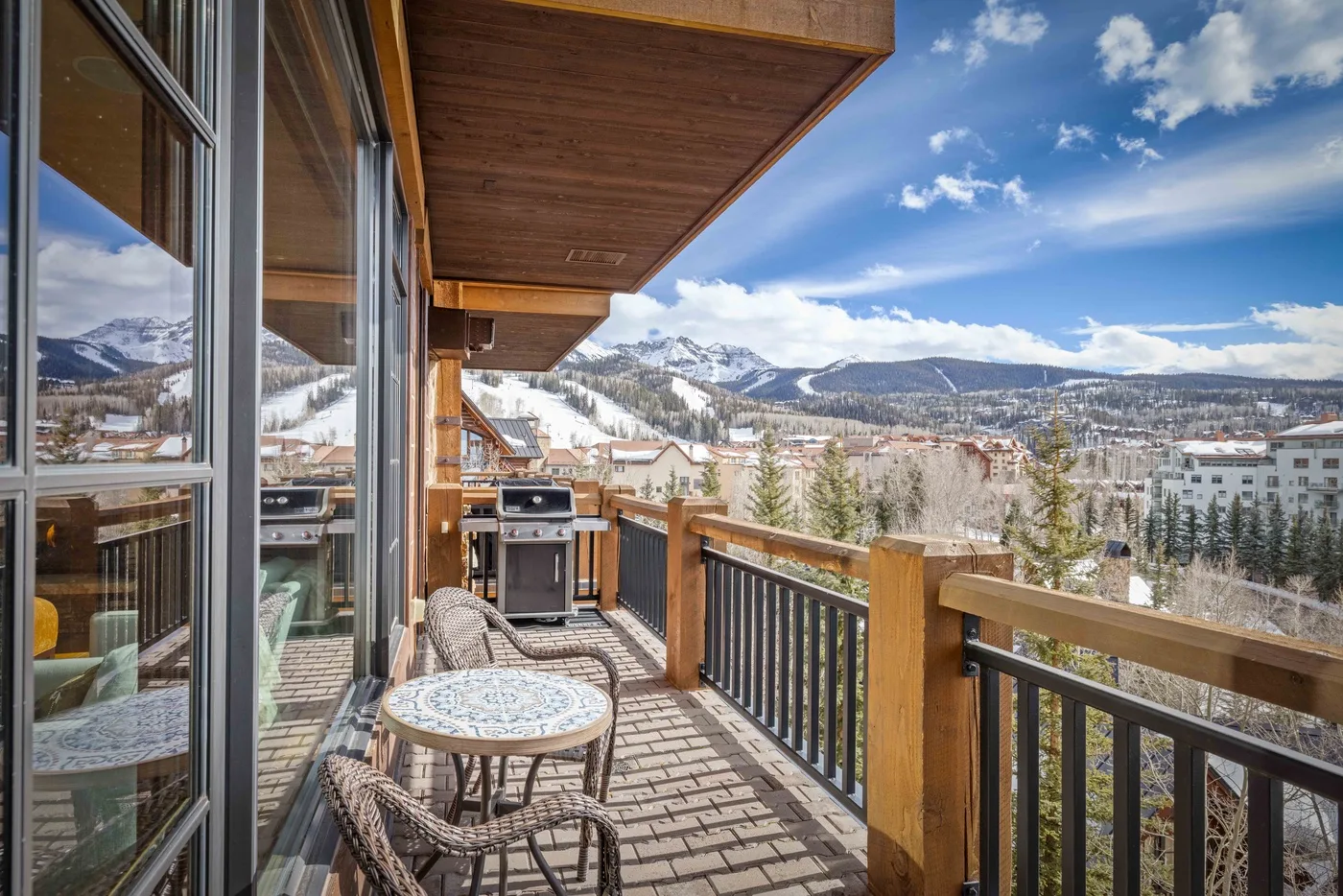 A wooden balcony with outdoor seating overlooks a town and snowy mountains.