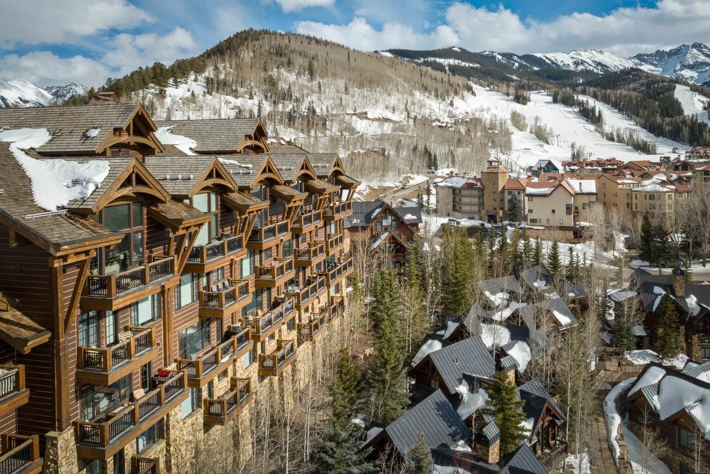 A mountain village with timber buildings sits below snow-covered slopes.