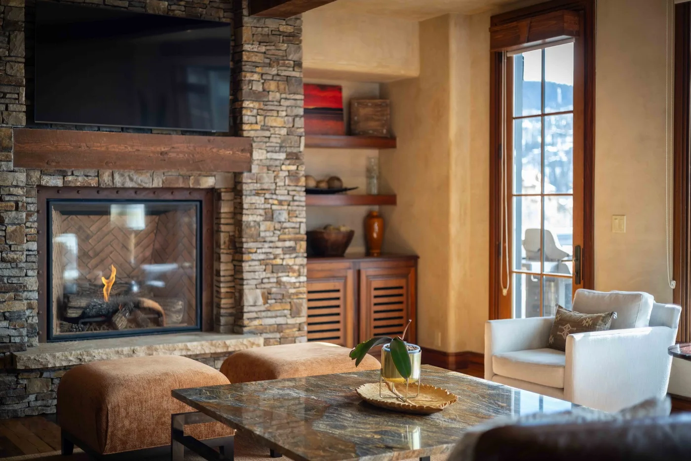 A living room with a stone fireplace, television, and seating by a tall window.
