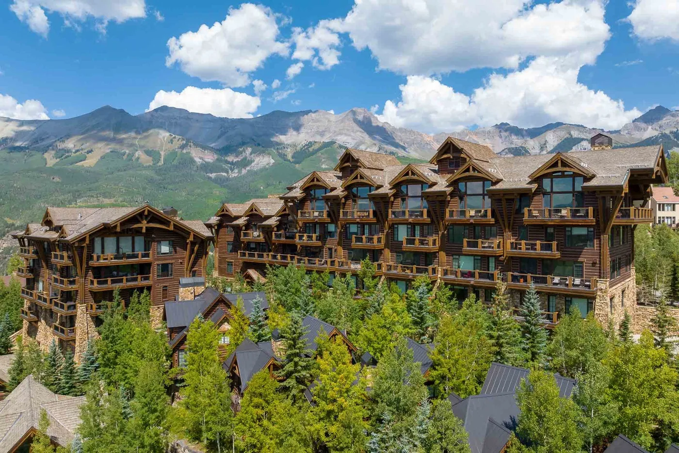 Un gran complejo de alojamientos de madera se alza entre árboles, frente a una cordillera.