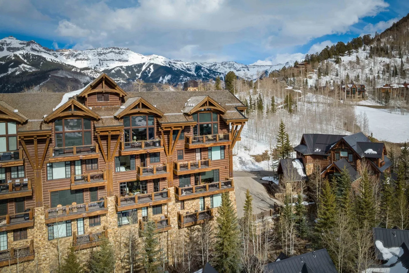 Mountain lodge buildings sit below a snowy hillside and distant peaks.