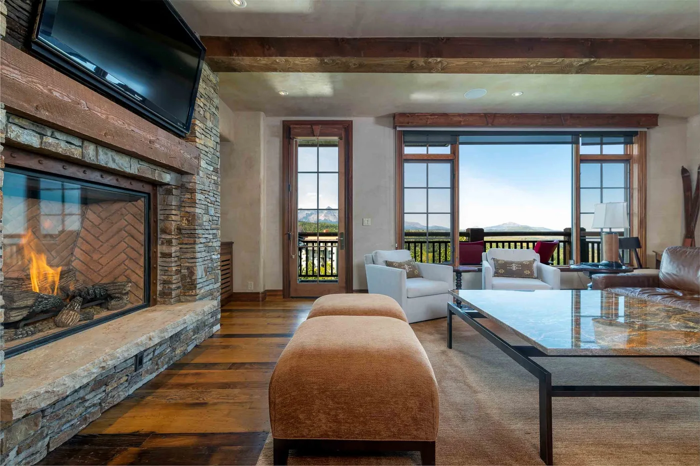 A living room with a stone fireplace, seating, and large windows overlooking a balcony.