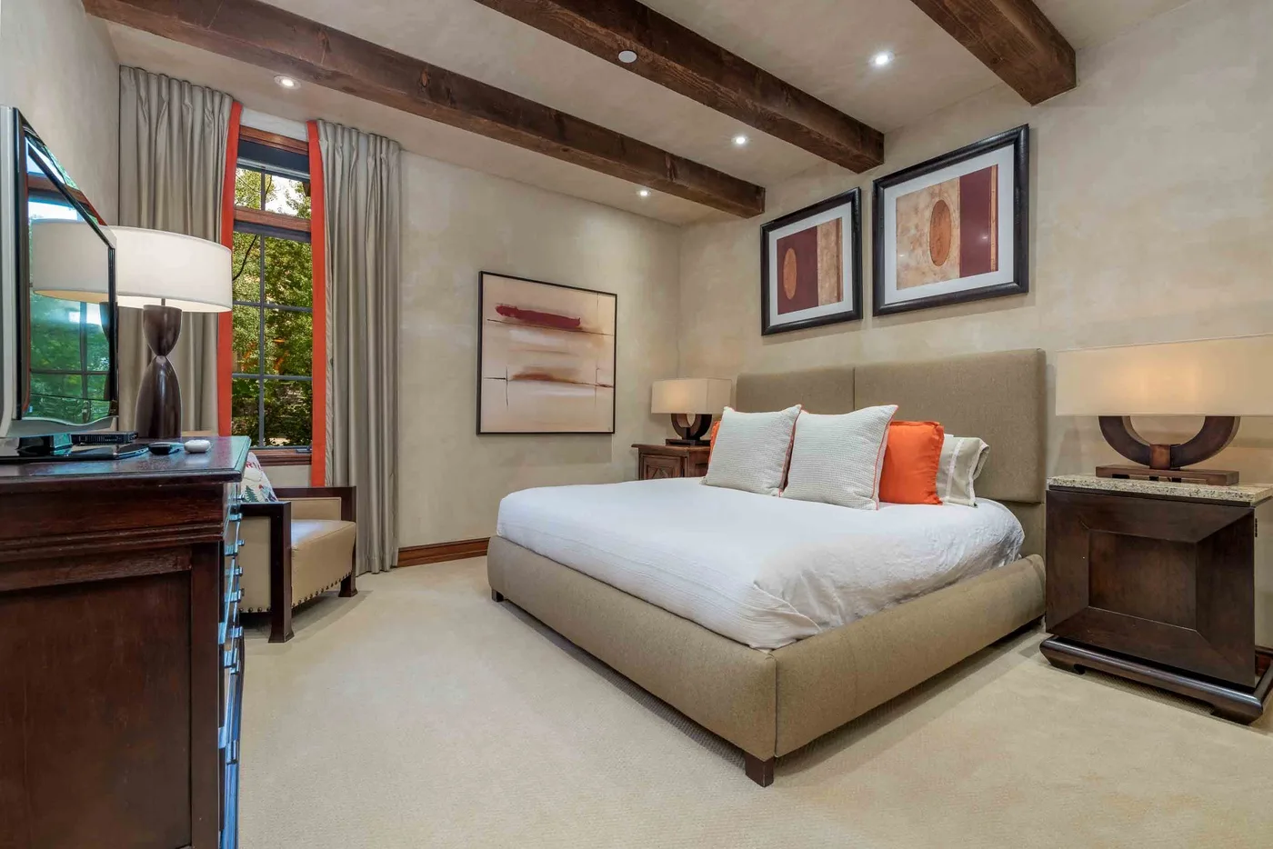 A bedroom with a neatly made bed, framed artwork, and exposed ceiling beams.