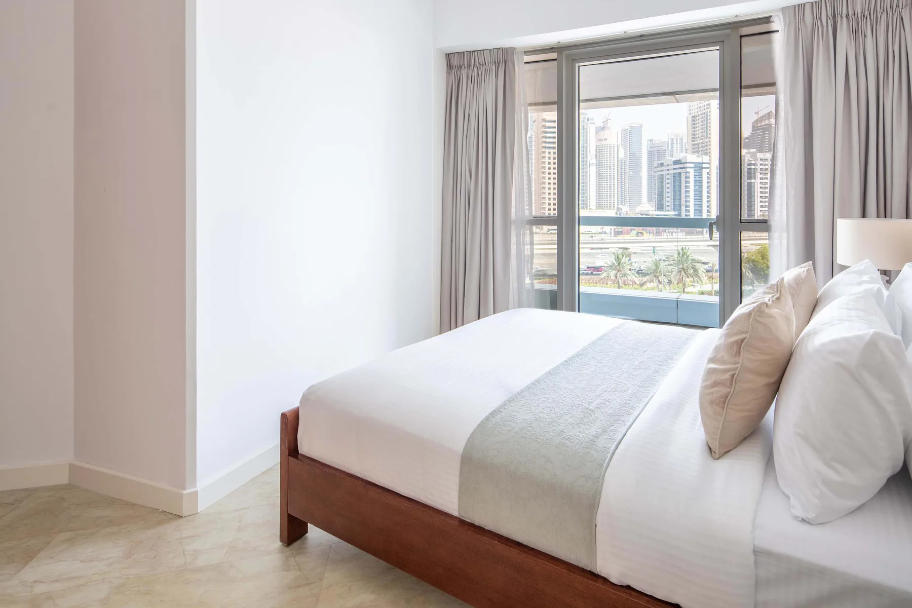 Un dormitorio luminoso con una cama bien hecha y grandes ventanas con vistas a edificios cercanos.