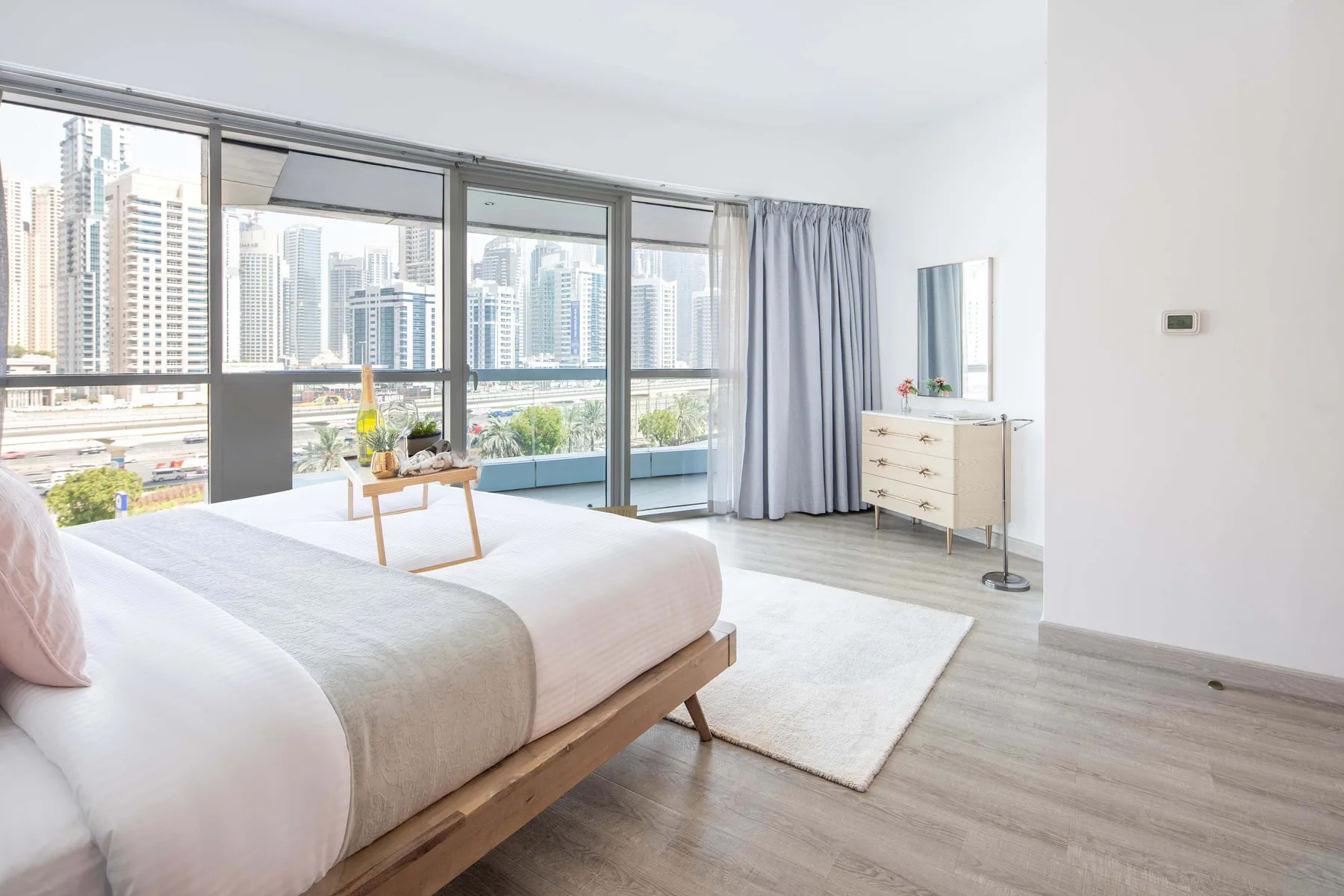 Un dormitorio luminoso con una cama, una cómoda y grandes ventanales con vistas a edificios de la ciudad.