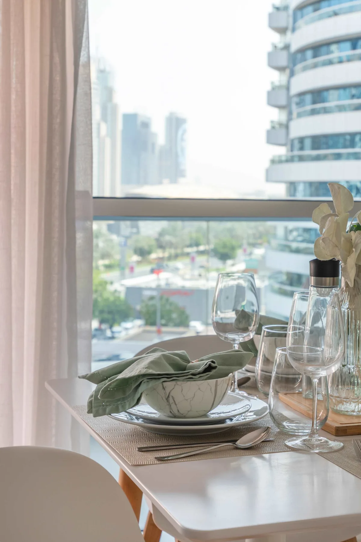 Una mesa de comedor preparada con platos está junto a una ventana con vista a edificios cercanos.