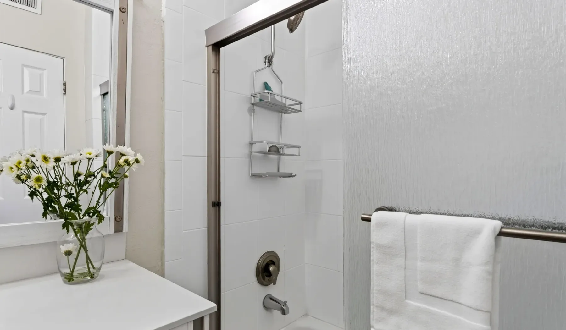 Un baño luminoso con ducha de vidrio, toallas blancas y flores sobre la encimera.
