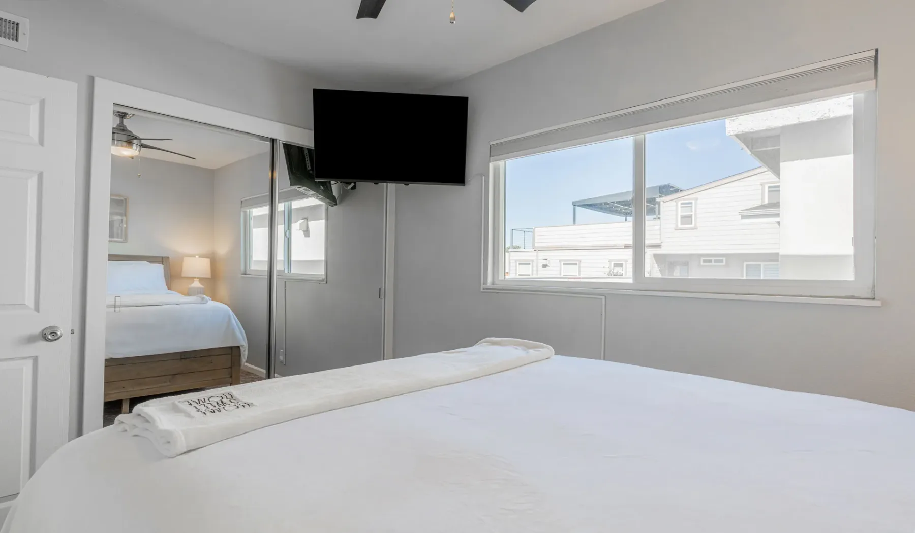 Un dormitorio luminoso con una cama grande, un televisor montado en la pared y una ventana amplia.