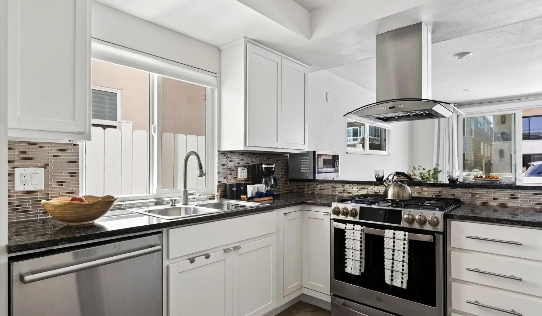 Una cocina luminosa con gabinetes blancos, encimeras oscuras y electrodomésticos de acero inoxidable.